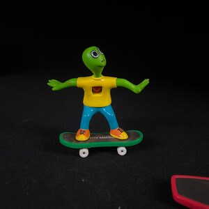 May include: A green alien figure with a yellow shirt and blue pants is riding a green and black toy skateboard with white wheels. The skateboard has the words "Toy Machine" printed on it.