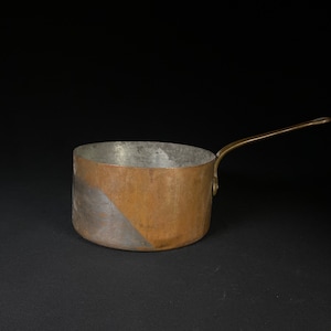 May include: A copper saucepan with a brass handle. The saucepan has a silver-colored interior and a copper exterior. The copper is tarnished and has a patina.