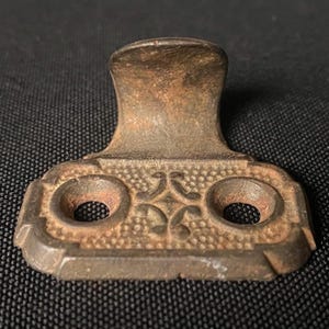 May include: A rusty cast iron window latch with a decorative floral design and two holes. The latch is in the shape of a rectangle with a curved top.