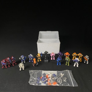May include: A collection of miniature Halo action figures in various colors, including red, blue, orange, and yellow. Some figures are assembled, while others are in a clear plastic bag. A white box is in the background.