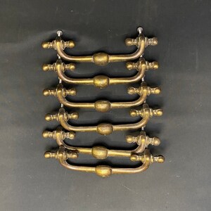 May include: A set of eight antique brass drawer pulls with ornate details. The pulls are arranged in a stack, with the top pull facing the viewer.