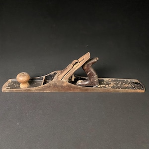 May include: A vintage wooden hand plane with a dark brown finish. The plane features a wooden knob and handle, and a metal blade. The tool shows signs of wear, with paint chipping off the surface. The plane is designed for woodworking.