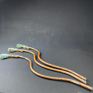 May include: Three antique copper-colored metal candle holders with a green patina. Each holder has a curved stem and a decorative leaf-shaped top.