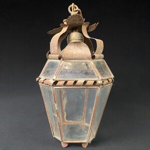 May include: A rustic, antique lantern with a glass body and a metal frame. The lantern has a decorative crown-like top and a chain for hanging. The metal is rusted and shows signs of age.