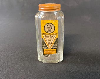 May include: Vintage Sta-Bac Curl Set bottle with a gold-colored lid and a yellow label. The label features a portrait of a woman and text indicating the product is for water and finger waving. The clear glass bottle has a rectangular shape.