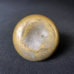 May include: A round, gold-colored metal object with a smooth, slightly textured surface. The object has a circular indentation in the center.