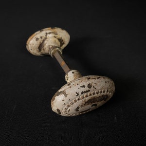 May include: White porcelain door knob with a decorative border and a metal spindle. The knob has a worn, aged look with brown spots.