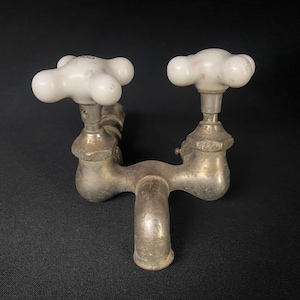 May include: Antique double faucet with porcelain knobs. The faucet is made of metal and has a silver finish. The knobs are white and have a cross-shaped design.