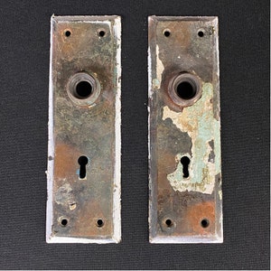 May include: Two antique door plates with a keyhole and a round knob. The plates are made of metal and have a worn, distressed finish with chipped paint in shades of green, blue, and brown.