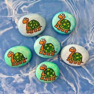 May include: Five painted rocks with a turtle design. The turtles are painted in shades of green, brown, and orange. The rocks are on a blue and white background.
