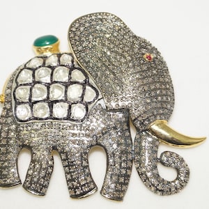 May include: An elephant-shaped brooch covered in small pavé-set stones with a dark metallic finish. The elephant features a gold tusk, a small red stone eye, and a saddle blanket adorned with larger white stones and a single green cabochon.