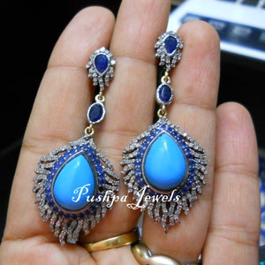 Peut inclure: Une paire de boucles d'oreilles pendantes avec une pierre turquoise centrale en forme de poire entourée de petits saphirs bleus et de diamants pavés. Le design inclut un saphir ovale et un saphir en forme de poire. Le texte Pushpa Jewels est visible.
