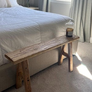 May include: A rustic wooden bench with a natural wood finish. The bench is made of reclaimed wood and has a simple, minimalist design. It is placed next to a bed in a bedroom with a white duvet and a window with white curtains.