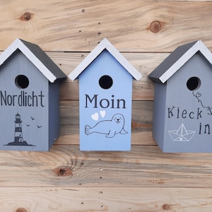 Op de afbeelding: Drie houten vogelhuisjes geschilderd in tinten blauw en grijs. De vogelhuisjes hebben verschillende ontwerpen, waaronder een vuurtoren, een zeehond en een zeilboot. De vogelhuisjes zijn gelabeld met "Nordlicht", "Moin" en "Kiek in".