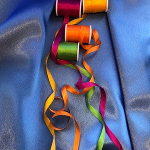 May include: Four spools of colorful thread with matching ribbons in orange, green, purple, and yellow. The ribbons are arranged in a loose, flowing pattern on a blue background.