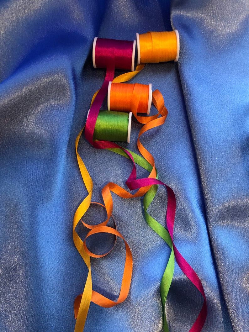 May include: Four spools of colorful thread with matching ribbons in orange, green, purple, and yellow. The ribbons are arranged in a loose, flowing pattern on a blue background.