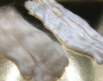 May include: Close-up of two piles of raw, undyed silk fibers. One pile is a loose, fluffy mass, while the other is composed of several bundles of long, wavy strands. The fibers are a creamy off-white color.