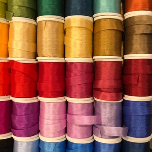 May include: A collection of spools of colorful silk ribbon, arranged in rows. The ribbon is wound around white spools with a white plastic cap on each end. The ribbon colors include green, blue, gold, red, purple, pink, light blue, and dark blue.