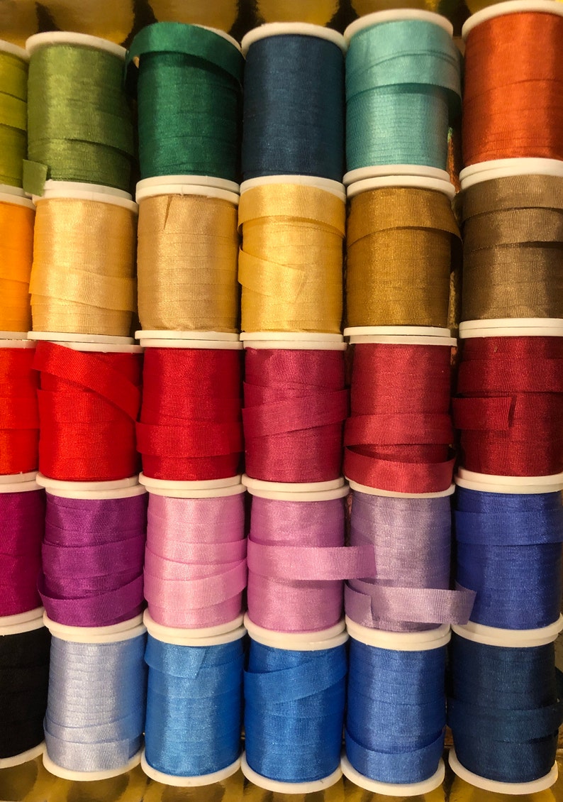 May include: A collection of spools of colorful silk ribbon, arranged in rows. The ribbon is wound around white spools with a white plastic cap on each end. The ribbon colors include green, blue, gold, red, purple, pink, light blue, and dark blue.