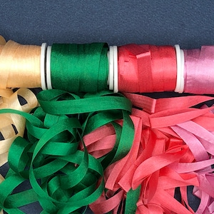 May include: Four spools of ribbon in various colors, including beige, green, red, and pink. The ribbon is unraveled and spread out on a dark blue surface.