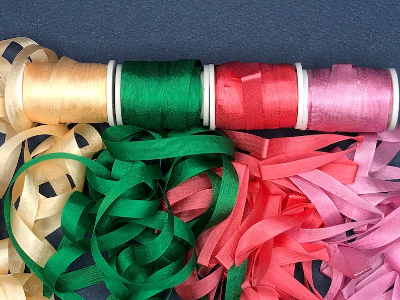May include: Four spools of ribbon in various colors, including beige, green, red, and pink. The ribbon is unraveled and spread out on a dark blue surface.
