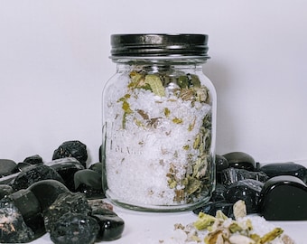 May include: A glass jar with a black lid filled with white bath salts and green herbs. The jar is sitting on a white surface surrounded by black stones.