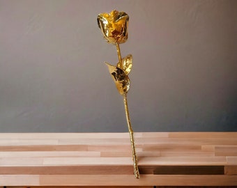 May include: A single golden rose with a long stem and leaves. The rose is on a wooden surface.