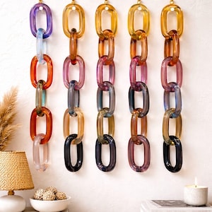 Könnte beinhalten: Wanddekoration mit fünf vertikalen Ketten aus farbigen, oval geformten Gliedern. Die Ketten bestehen aus durchscheinendem Glas in Lila, Orange, Rosa und Bernstein. Jede Kette ist mit einer goldfarbenen Halterung an der Wand befestigt.