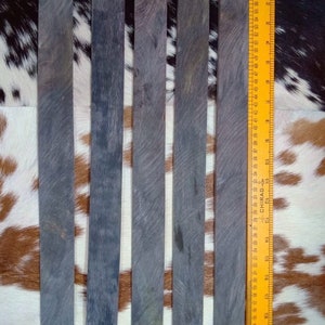 Puede incluir: Cinco listones de madera gris oscuro, cada uno de aproximadamente 30 cm de largo, están dispuestos uno al lado del otro sobre una piel de vaca marrón y blanca. Una regla amarilla es visible en el lado derecho de la imagen.