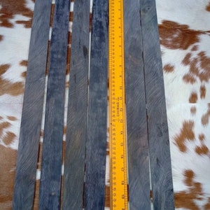 May include: Six dark gray wooden dowels, each approximately 21 inches long, are arranged side-by-side on a brown and white patterned surface. A yellow ruler is placed next to the dowels for scale.