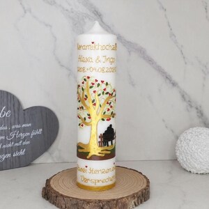 Könnte beinhalten: Eine weiße zylindrische Stumpenkerze mit einer goldenen Baumillustration mit roten herzförmigen Blättern, einer schwarzen Silhouette von zwei Personen auf einer Bank und dem goldenen Text Keramikhochzeit Alexa & Ingo 04.08.2020 sowie Zwei Herzen ein Versprechen.