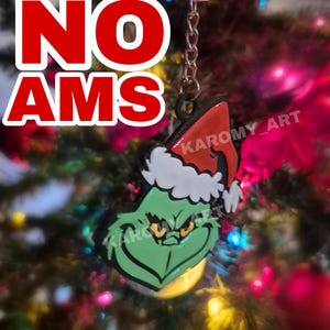 Puede incluir: Un llavero con temática de Grinch con una cara verde y una expresión enfadada, que lleva un gorro de Papá Noel rojo y blanco. El llavero tiene una cadena plateada y el texto "NO AMS" en rojo. También es visible el nombre del artista, "KAROMY_ART".