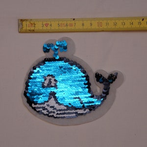 Könnte beinhalten: Eine blaue und silberne Pailletten-Wal-Applikation. Der Wal blickt nach rechts und hat eine kleine Schwanzflosse. Die Applikation befindet sich auf einem weißen Hintergrund.