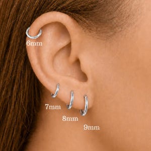May include: Silver hoop earrings in 6mm, 7mm, 8mm, and 9mm sizes are displayed on an ear. The small, round earrings are shiny. The image shows the earrings in place on the ear.