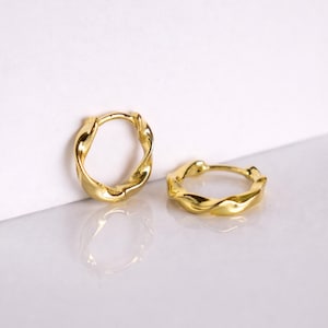 May include: A pair of gold-coloured hoop earrings with a twisted design. The earrings are presented on a white surface with a white backdrop. The earrings have a shiny, reflective surface.