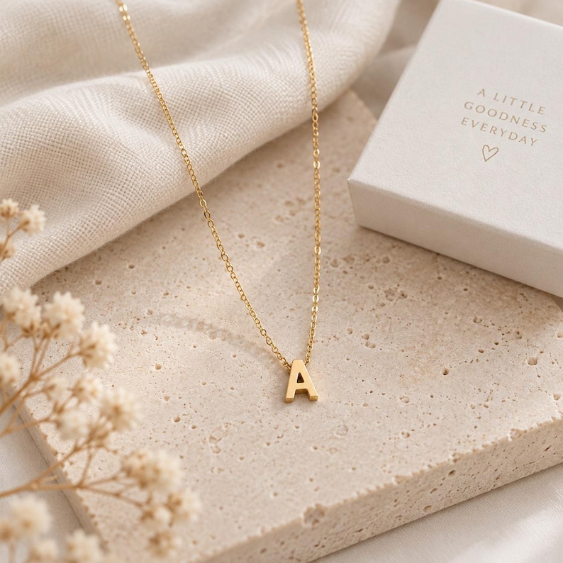 May include: A gold-toned necklace featuring a delicate chain and a single letter A pendant. The necklace is presented alongside a white gift box with the text A LITTLE GOODNESS EVERYDAY and a small heart symbol printed on the top surface.