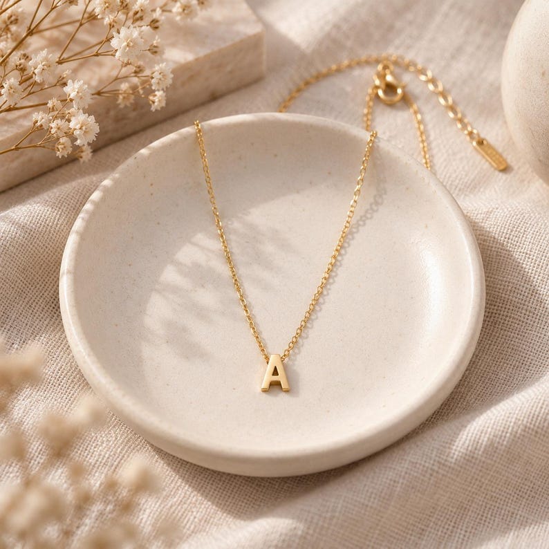 May include: A gold-toned necklace featuring a delicate chain and a single capital letter A pendant. The necklace includes an adjustable chain with a small metal tag at the end, showcasing a polished finish that reflects light across the surface.