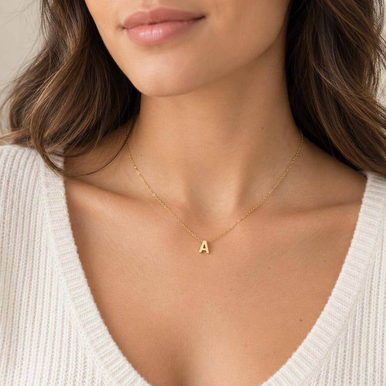 May include: A gold-coloured necklace featuring a delicate chain and a single, centred capital letter A pendant. The metal has a polished finish, and the pendant hangs centrally from the thin, lightweight chain, resting against the collarbone area.