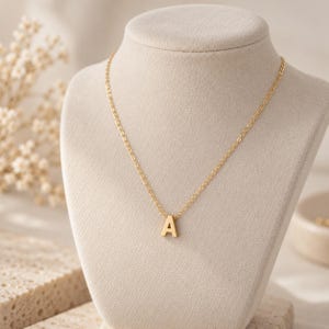 May include: A gold-toned necklace featuring a delicate chain and a single capital letter A pendant. The pendant is centered on the chain, which is displayed on a neutral-colored bust. The metal has a smooth, polished finish throughout the piece.