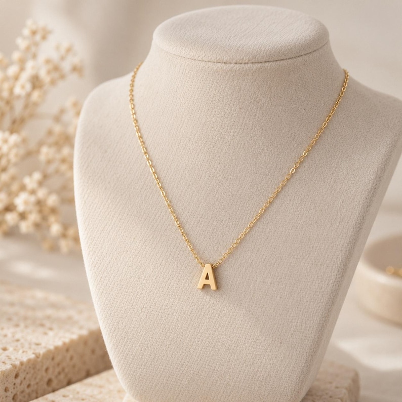 May include: A gold-toned necklace featuring a delicate chain and a single capital letter A pendant. The pendant is centered on the chain, which is displayed on a neutral-colored bust. The metal has a smooth, polished finish throughout the piece.