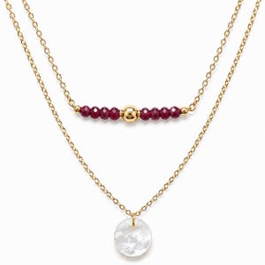 Puede incluir: Un collar de doble cadena dorada que presenta una hebra superior con una cuenta dorada central flanqueada por cuentas facetadas de color rojo oscuro y una hebra inferior con un colgante de disco blanco iridiscente en el centro.