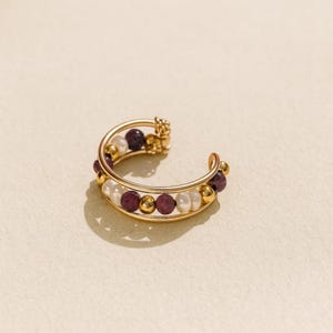 Peut inclure: Un ear cuff doré avec un design incurvé. Il est orné de petites perles rondes blanches, rouge foncé et dorées. Le cuff est conçu pour s'enrouler autour de l'oreille, un bijou élégant.