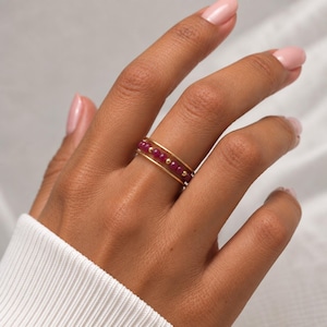 May include: A gold ring featuring a central row of small, round, deep pink beads interspersed with tiny gold spheres, framed by two thin, smooth gold bands on either side, creating a layered appearance on the finger.