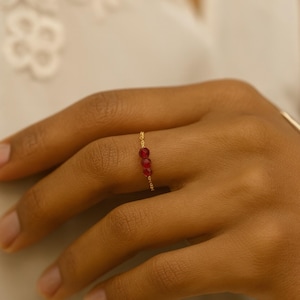 Peut inclure: Une fine bague en or ornée de trois petites pierres précieuses rouges vives. La bague a un design fin et minimaliste, avec les pierres précieuses rapprochées. La bague est présentée sur une main, soulignant son design élégant et son contraste de couleurs.