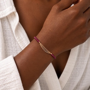 Può includere: Un bracciale caratterizzato da un filo di piccole perline rotonde color rosa intenso, interrotto da una sezione centrale composta da tre sottili e delicate catenelle in metallo dorato che pendono leggermente, unite alle perline da due sfere dorate.