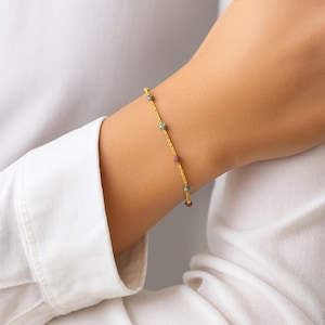 May include: A delicate gold bracelet featuring small, colorful beads. The bracelet is strung on a fine gold chain and is worn on the wrist. The beads are in shades of green, red, and brown. The bracelet is a piece of jewelry.