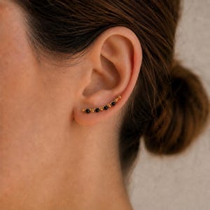 Peut inclure: Une boucle d'oreille grimpante dorée composée d'une rangée incurvée de petites perles noires rondes alternant avec de minuscules sphères dorées. Le bijou suit la courbe naturelle du lobe de l'oreille, formant une ligne structurée et contrastée.