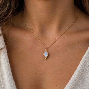 Può includere: Una collana a catena dorata con un ciondolo in pietra di luna a forma di goccia. Il ciondolo è incastonato in una montatura dorata con tre piccole sfere dorate alla base. La collana poggia sul collo, mettendo in risalto il riflesso blu iridescente.