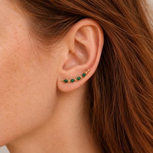 May include: A gold ear climber earring featuring a curved row of five small, round, deep green stones alternating with tiny gold beads. The earring follows the natural curve of the earlobe, creating a delicate and structured line of colour.