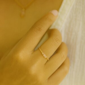 Peut inclure: Une fine bague en or ornée de trois petites pierres roses pâles. La bague a un fin anneau torsadé et est portée au doigt. L'arrière-plan est d'une tonalité douce et chaude, mettant en valeur le bijou.